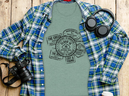 Rugged Wanderlust Passport Stamp Graphic Wilderness Shirt: Green t-shirt with passport stamp graphic, worn under a plaid shirt, with headphones and camera.