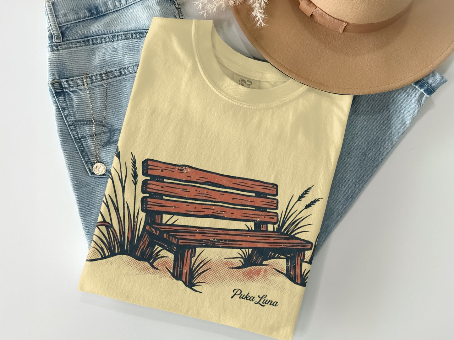 Cream t-shirt with a wood bench and grass illustration and "Puka Luna" text.