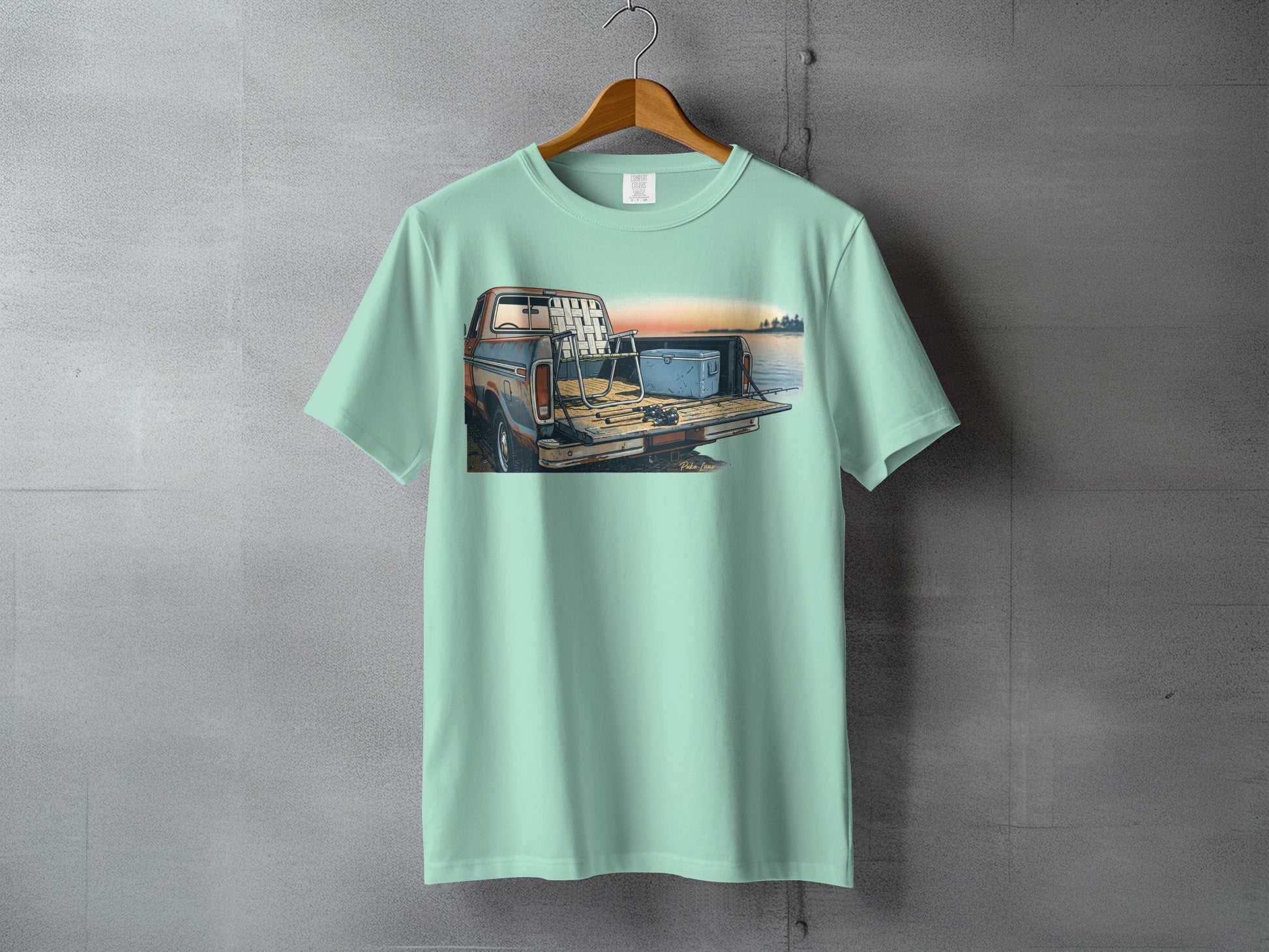 Mint green t-shirt with graphic of a truck, fishing rods, and cooler by water at sunset.