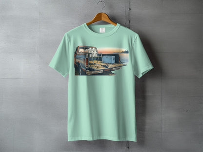 Mint green t-shirt with graphic of a truck, fishing rods, and cooler by water at sunset.