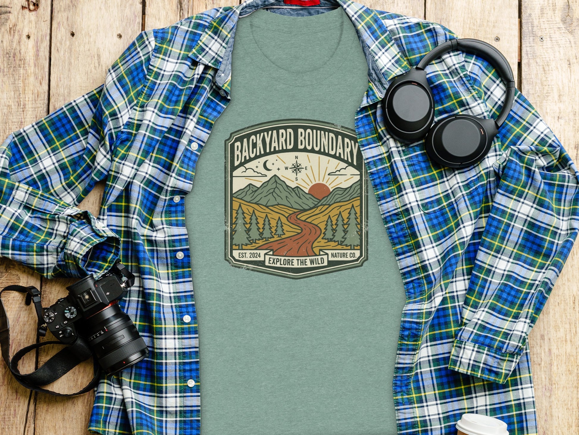 Rugged Backyard Boundary: Explore The Wild Wilderness Shirt: Green t-shirt with "Backyard Boundary Explore the Wild" logo, set on a wooden table.