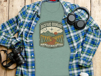 Rugged Backyard Boundary: Explore The Wild Wilderness Shirt: Green t-shirt with "Backyard Boundary Explore the Wild" logo, set on a wooden table.