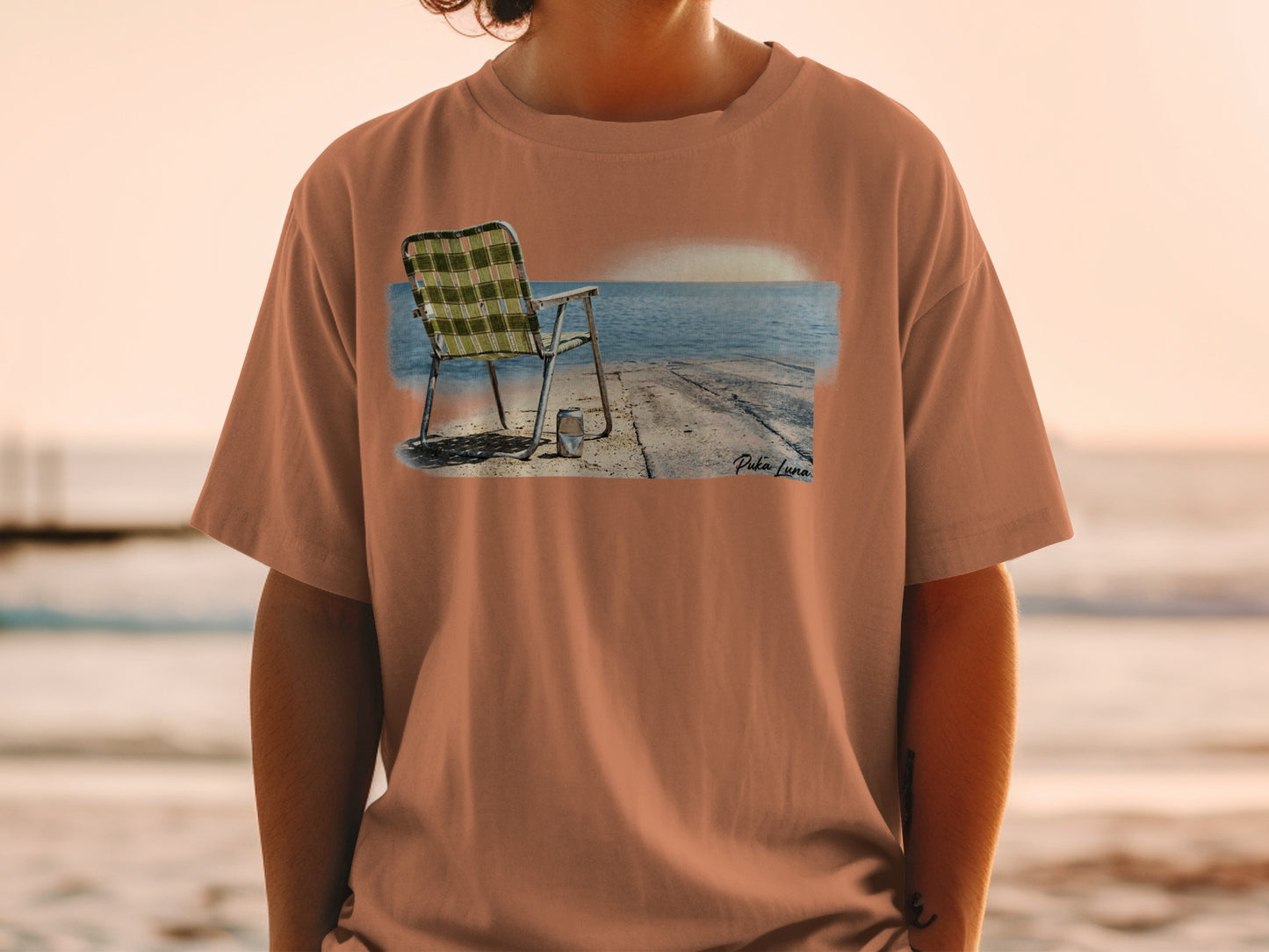 T-shirt featuring a printed graphic of a beach chair and cooler by the ocean.