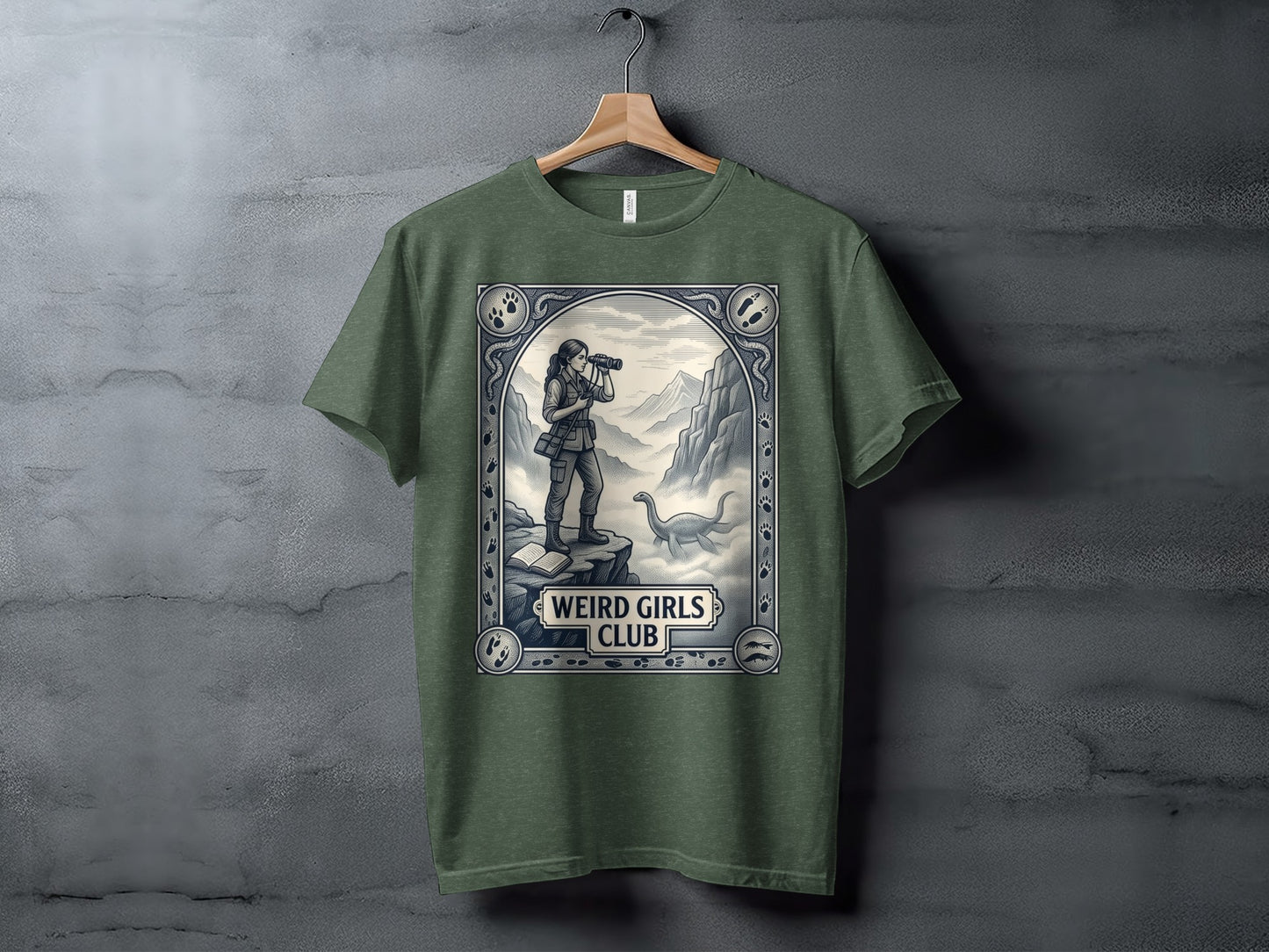 Olive green t-shirt featuring a "Weird Girls Club" design with a woman observing a dinosaur.
