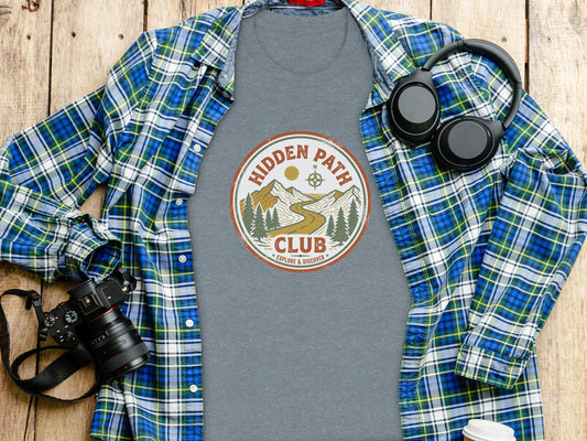 Rugged Hidden Path Club Explorer Wilderness Shirt: Gray t-shirt with "Hidden Path Club" circular logo, worn under a blue and green plaid shirt with headphones.