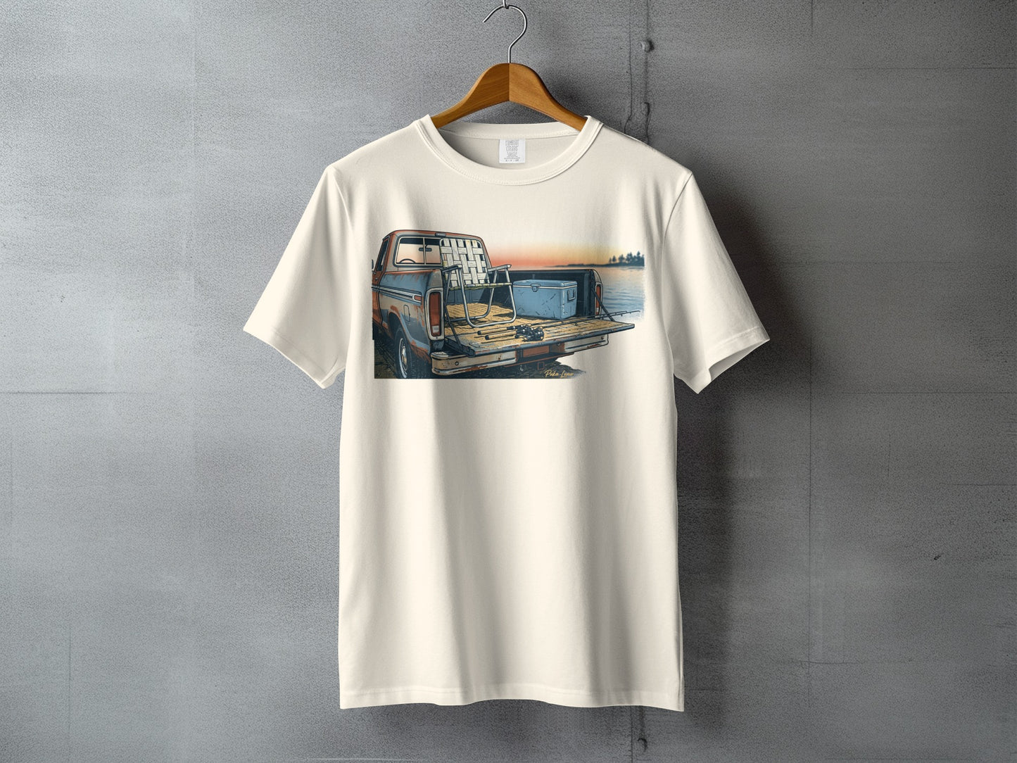 Cream t-shirt with illustration of a pickup truck bed with fishing gear at sunset.