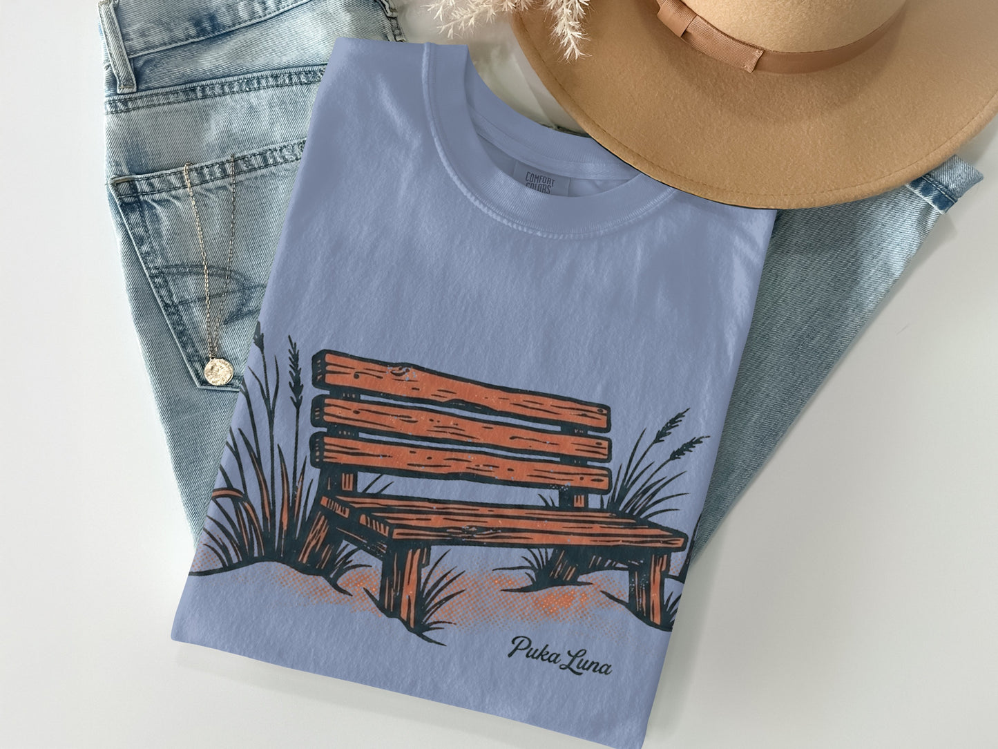 Light blue t-shirt with a graphic of a wooden bench and reeds, and the text "Puka Luna".