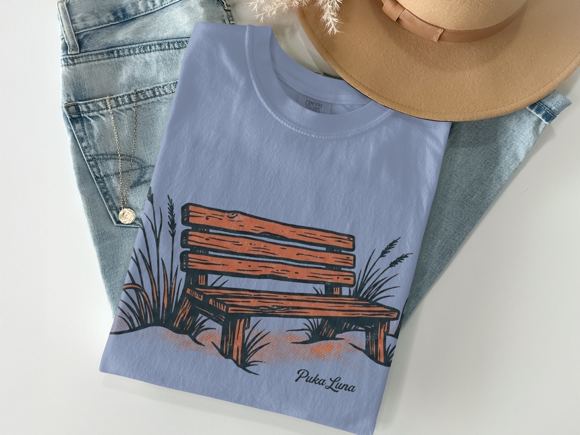 Light blue t-shirt with a graphic of a wooden bench and reeds, and the text "Puka Luna".