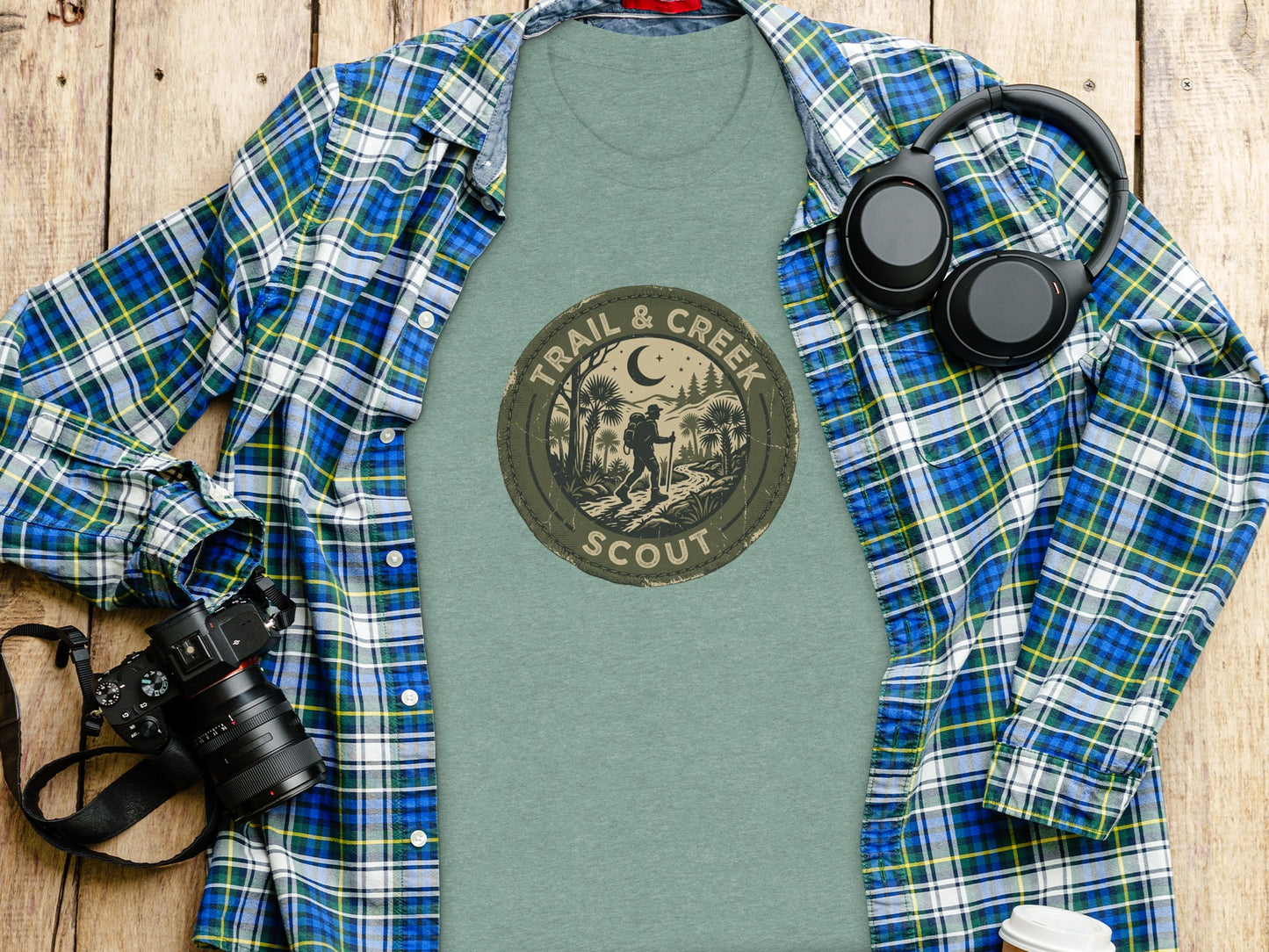 Heritage Trail & Creek Scout Adventure Nomad Tee: Green t-shirt with "Trail & Scout" circular logo displayed on wooden surface with headphones and camera.