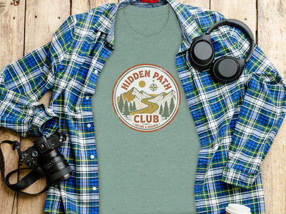 Rugged Hidden Path Club Explorer Wilderness Shirt: Green t-shirt with "Hidden Path Club" logo, camera, and headphones on wooden surface.