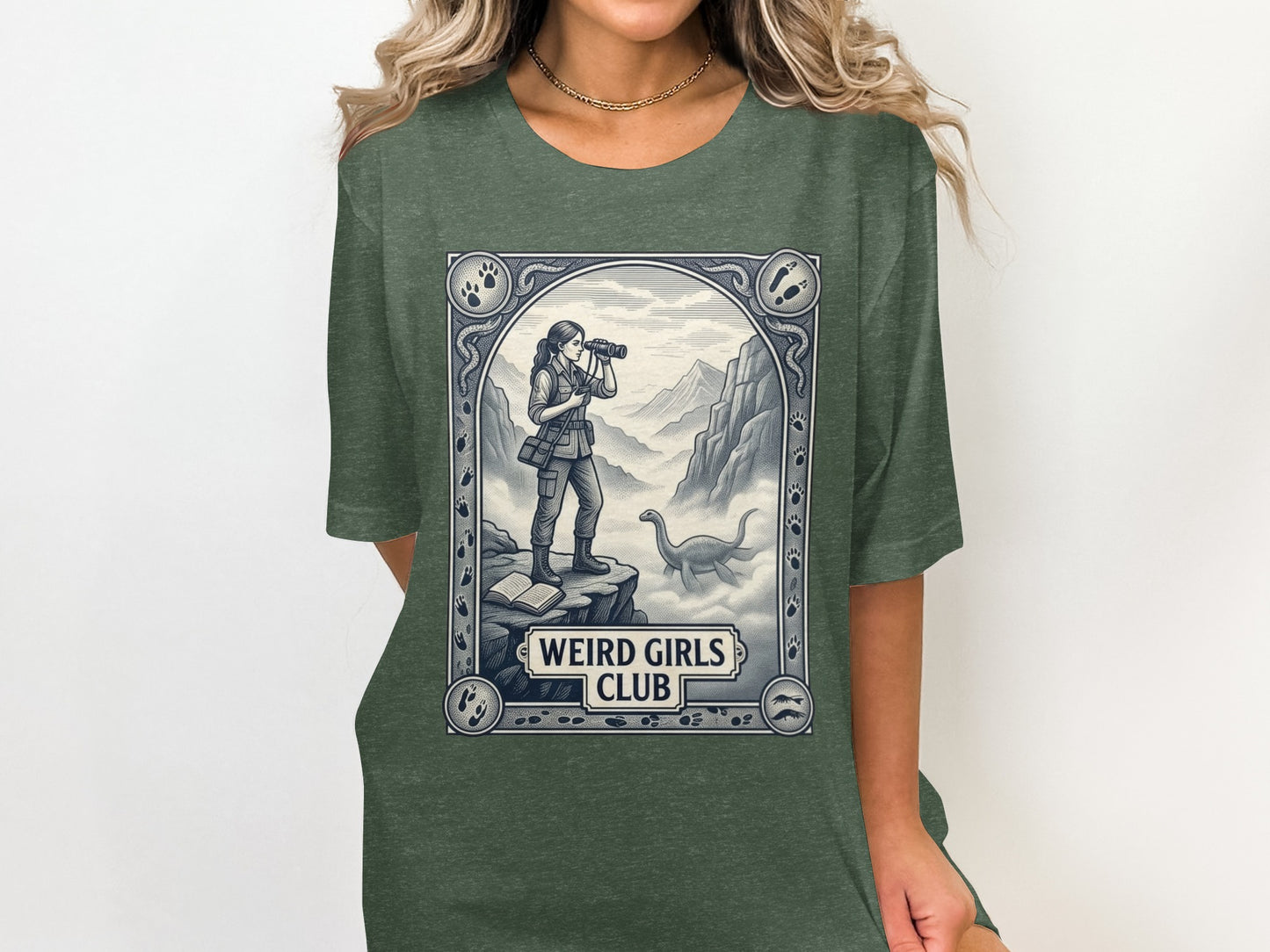T-shirt graphic of a woman with binoculars on a cliff overlooking mountains and a dinosaur, with "WEIRD GIRLS CLUB" text.