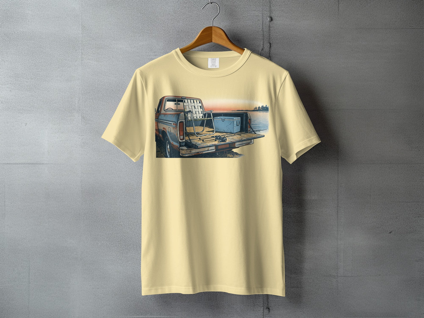 Cream t-shirt with illustration of a pickup truck bed with fishing gear by a lake.