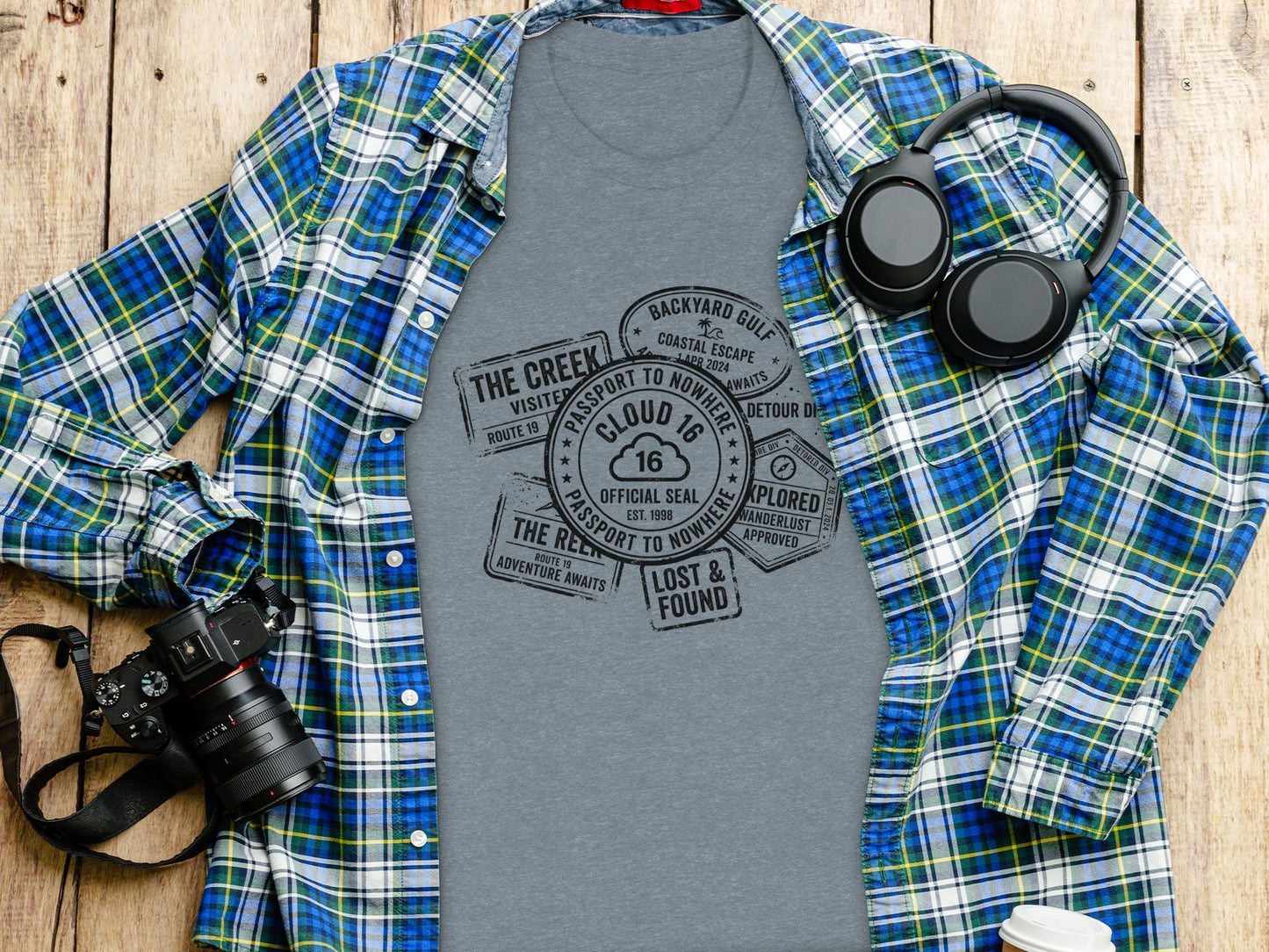 Rugged Wanderlust Passport Stamp Graphic Wilderness Shirt: Gray t-shirt with passport stamp graphic, plaid shirt, camera, and headphones.