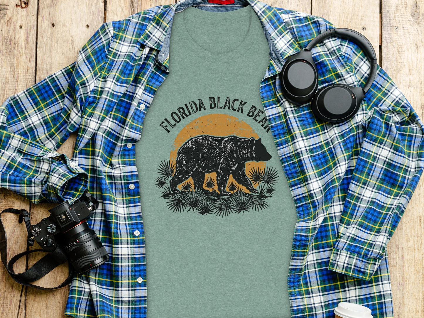 Heritage Florida Bear Trails Classic Nomad Tee: Green t-shirt with a Florida Black Bear graphic, headphones, and camera on a wooden surface.