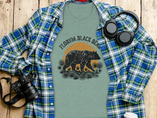 Heritage Florida Bear Trails Classic Nomad Tee: Green t-shirt with a Florida Black Bear graphic, headphones, and camera on a wooden surface.