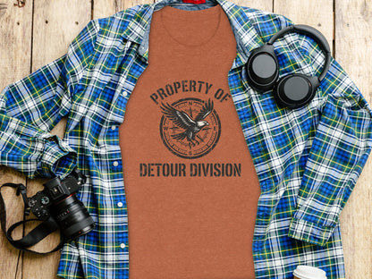 Rugged Detour Division Compass Eagle Tee Wilderness Shirt: Brown t-shirt with "Property of Detour Division" and eagle graphic, laid out with headphones and camera.