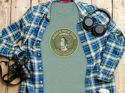 Hidden Path Club: Secret Entrance | Vintage Outdoor Apparel: Green t-shirt with "Hidden Path Secret Entrance" circular logo, laid flat with camera and headphones.